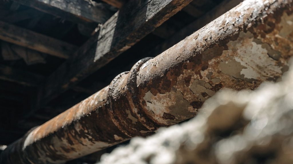 Old heavily corroded cast iron pipe exposed in a dim basement crawlspace, close inspection angle