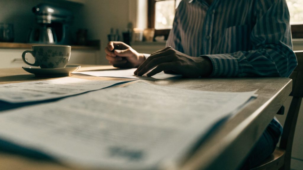 Person at a kitchen table reviewing paperwork with a coffee cup, blurred out-of-focus document, cozy home lighting, torso and hands only