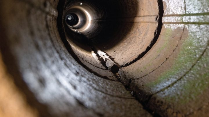 Sewer inspection camera view looking down a dim clay pipe interior toward a cracked pipe joint, tunnel lighting, documentary style
