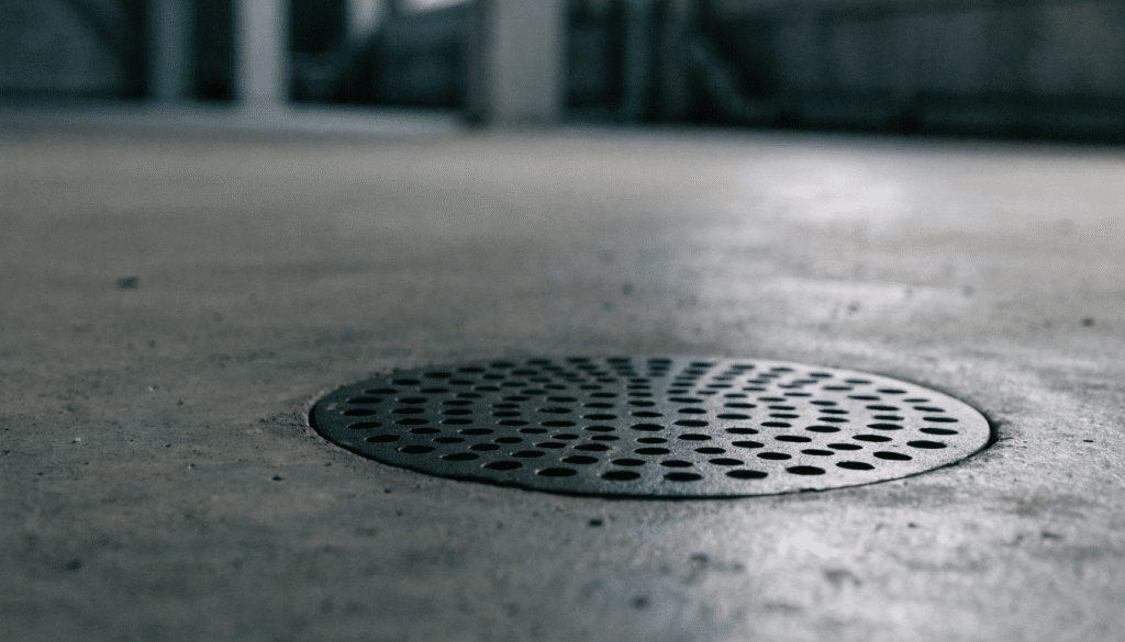 A round cast iron floor drain grate set into a bare basement concrete floor