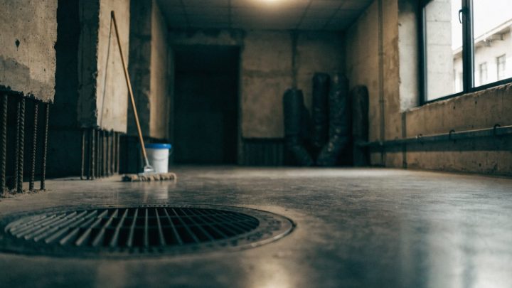 Unfinished concrete basement utility room with a floor drain, mop and bucket nearby, moody overhead light, clean and dry