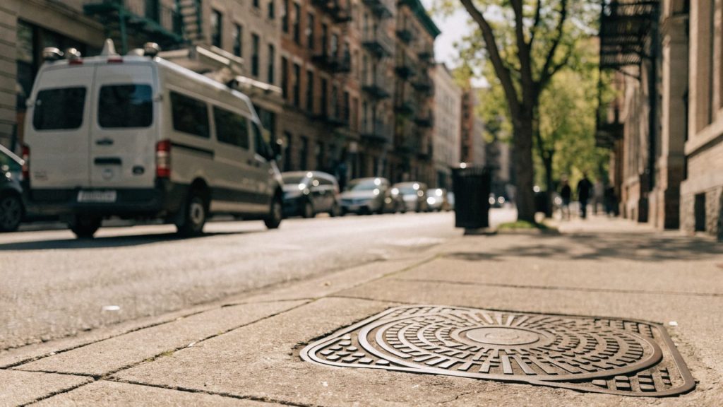 Residential street with a cast iron manhole cover and a utility inspection van parked at the curb, city sidewalk, daytime