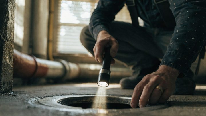 Plumber crouched at a floor drain in a basement utility room with a flashlight, inspecting for a slow drain, torso and hands only