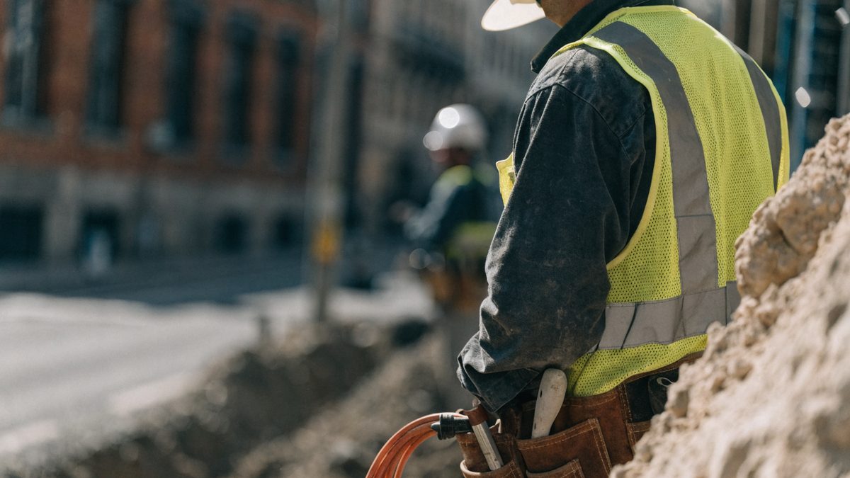 Utility worker in a safety vest and hard hat near an open city street excavation, torso only