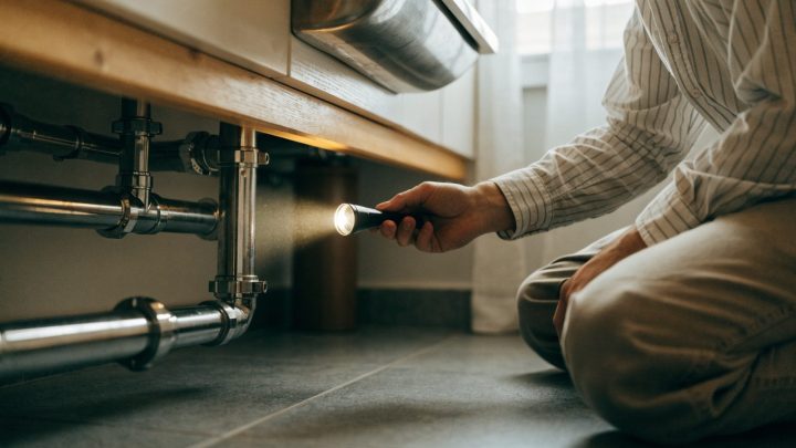 Person crouching to look underneath a bathroom sink cabinet with a flashlight, checking pipes, torso and hands only