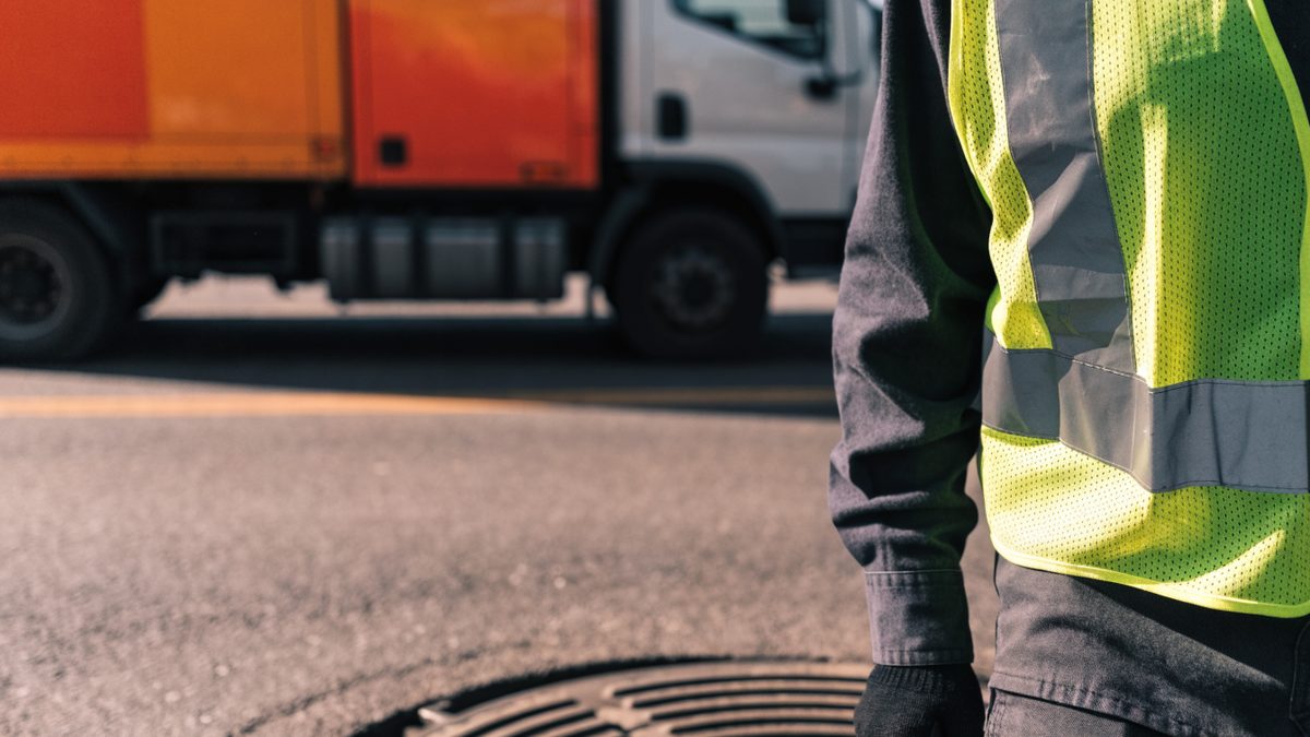 City utility worker in a safety vest near a street manhole, municipal truck blurred in background, torso only